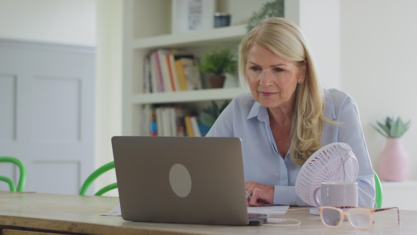 Menopausal mature woman working on laptop with connected fan at home having hot flush and fanning herself with documents - shot in slow motion Royalty-Free Stock Footage #1094252301
