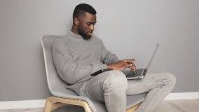 African-American male freelancer sitting in a chair at home against a gray wall and looking at a laptop screen typing on a keyboard with a phone on his foot, education and work online