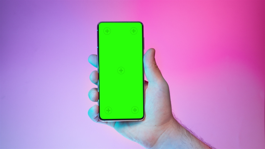 Mobile phone in hand. Holding smartphone with colorful red blue background. Green chromakey screen with markers. Neon lighting. Hand lifts phone up and holds it still. | Shutterstock HD Video #1094262979