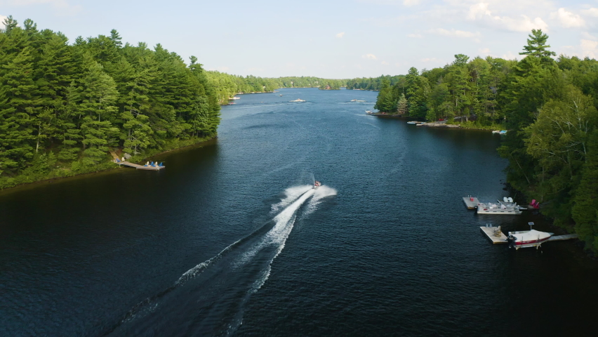 Jet ski drives down a beautiful blue river surrounded by trees. Aerial view revealing other boats in the distance.