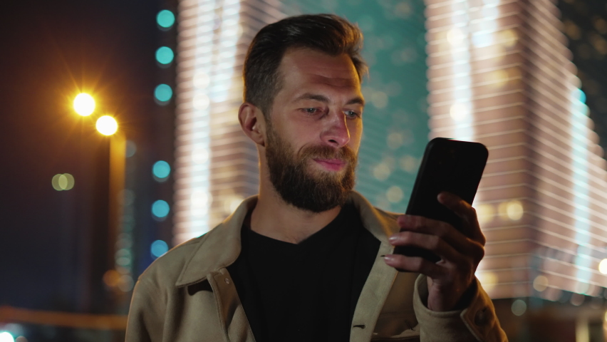 One Man with Phone in Hand at Dark Urban Street Sharing News. Person Walks in Neon Evening and Uses Smartphone Near Futuristic Illuminated Buildings. Electronic Information for Big Life of Traveler
