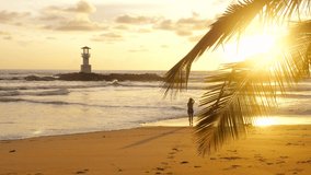 Alone woman walking on sunset beach with ocean waves and lighthouse on background. Cinematic video of travel girl enjoy summer vacation on the tropical sea. Holidays in Thailand