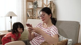 curious Chinese toddler baby getting close to her mother who is showing educational video on the mobile phone while sitting at leisure in the living room at home