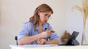 A young woman is sitting at a table and taking part in a video call. Using an app on a tablet for online learning and remote work, taking notes on paper.