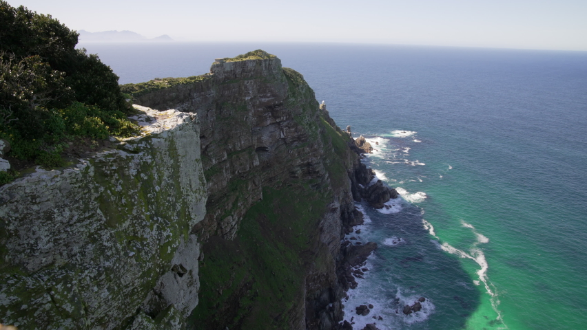 A dramatic view of the jagged rock-face and sheer cliffs of Cape Point, with the blue ocean and waves crashing below.