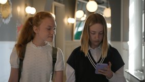 Schoolgirls use smartphone and talk walking in school corridor. Teenage girls with cellphone discuss news during break inside campus
