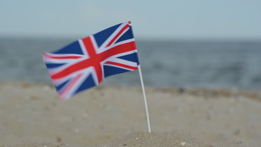 Great Britain flag on sandy beach on sea and waves background. British flag blowing in the wind.