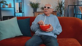 Excited elderly man on sofa eating popcorn and watching interesting tv serial, sport game, film, online social media movie content online at home. Senior grandfather enjoying domestic entertainment