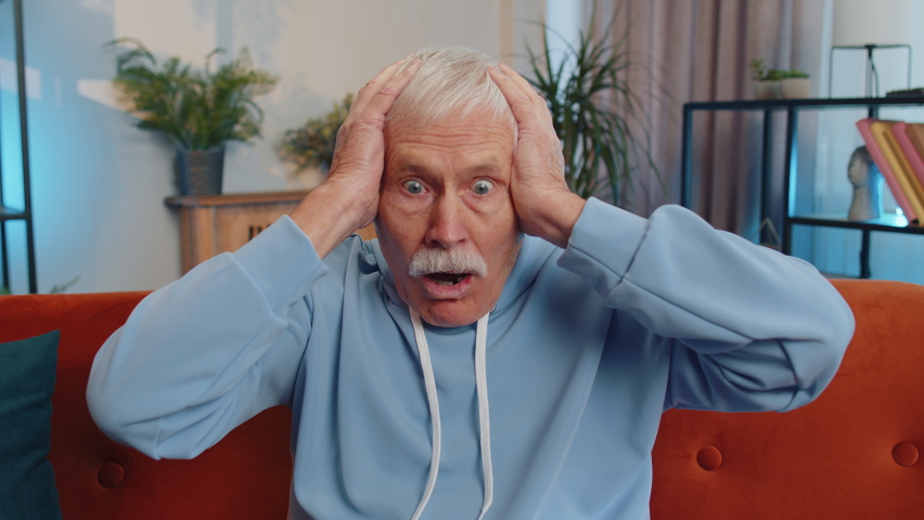 Oh my God, Wow. Mature elderly man surprised looking at camera with big eyes, shocked by sudden victory, good win news, celebrating at home. Excited amazed senior grandfather indoors in room on couch