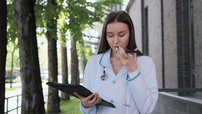 Professional female doctor standing near hospital holding documents recording voice message, using mobile phone virtual assistant. Doctor sends an online prescription to the patient during a pandemic.