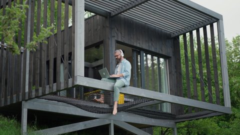 Стоковое видео: Man sitting on tiny house terrace working on laptop, listening to music