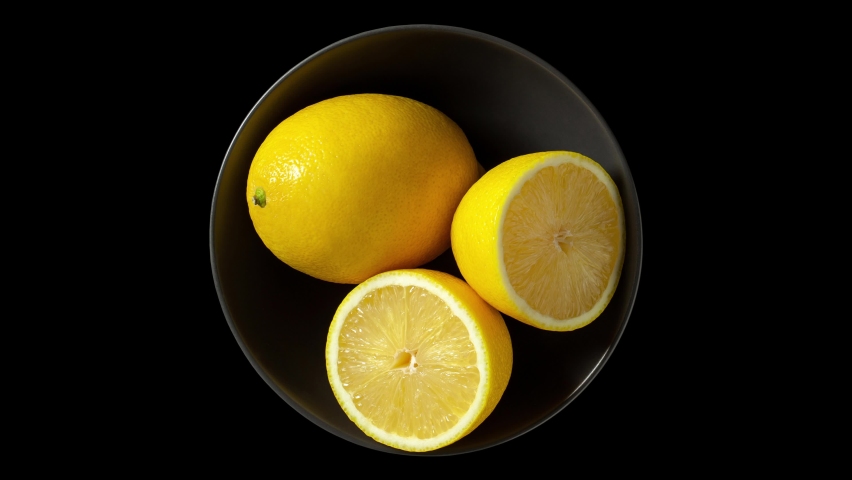 Lemons whole and cut in grey bowl, isolated on black background, rotating, turning, close-up macro, top view.
