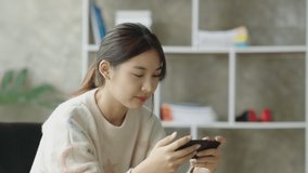 Beautiful and cute young Asian woman sitting on sofa and using smartphone happily and play game at her home.