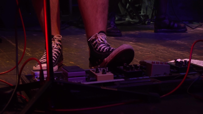 Foot in gumshoes pressing on loop pedal for electric guitar. Guitarist step on guitar effect pedal on rock concert in a club. Rock band with guitarists and drummer performing at concert in night club. Royalty-Free Stock Footage #1094307229