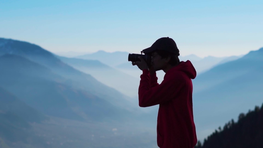 Silhouette shot of an Indian photographer taking photo in front of the foggy Himalayan mountains at Manali in Himachal Pradesh, India. Traveler taking photo of mountains. Man shoots landscape.  | Shutterstock HD Video #1094308865