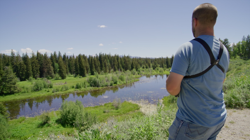 Male traveler looks through binoculars while exploring Grand Teton National Park. Picturesque wild trails along deep fir forest and grassy wetlands. High quality 4k footage