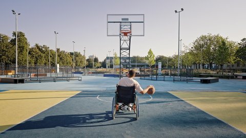 Disabled man in wheelchair improves basketball skills on outdoor court near city park. Guy practices to shoot ball into basket at sunset backside view Video stock