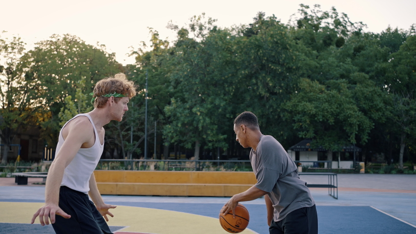 Friends compete playing basketball on outdoor city court against park with lush trees. Skilled black man makes goal shooting ball into basket slow motion
