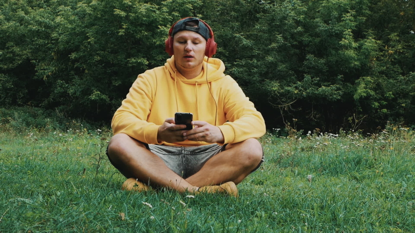 A young guy in headphones listening to music and humming his favorite song while sitting in nature. Man in over-ear headphones Royalty-Free Stock Footage #1094319751