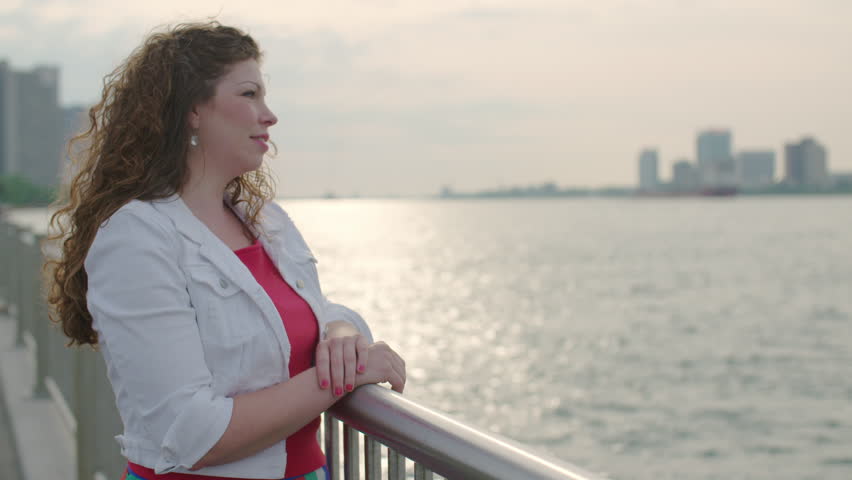 Attractive brunette watching the Detroit River.  Detroit and Windsor, Ontario, skylines visible in background.  Slow motion, recorded at 60fps in 4K.
