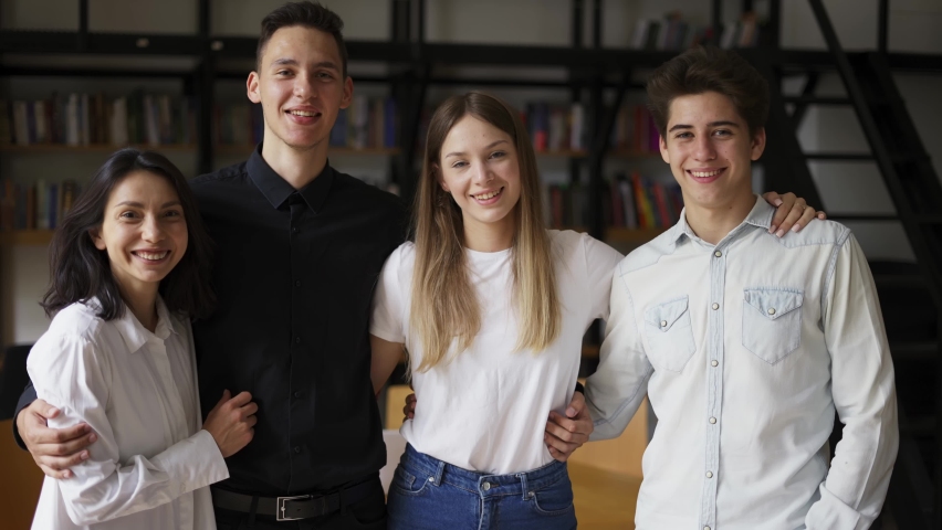 Zoom out portrait of four multiethnic young people standing together in group looking at camera posing at library or in modern office. Successful business team, students groupmates friends concept
