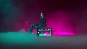 Female model, actress filming in a studio filled with smoke and neon lights background. Young woman in a hat sitting on a playback in the middle of the room. High quality 4k footage