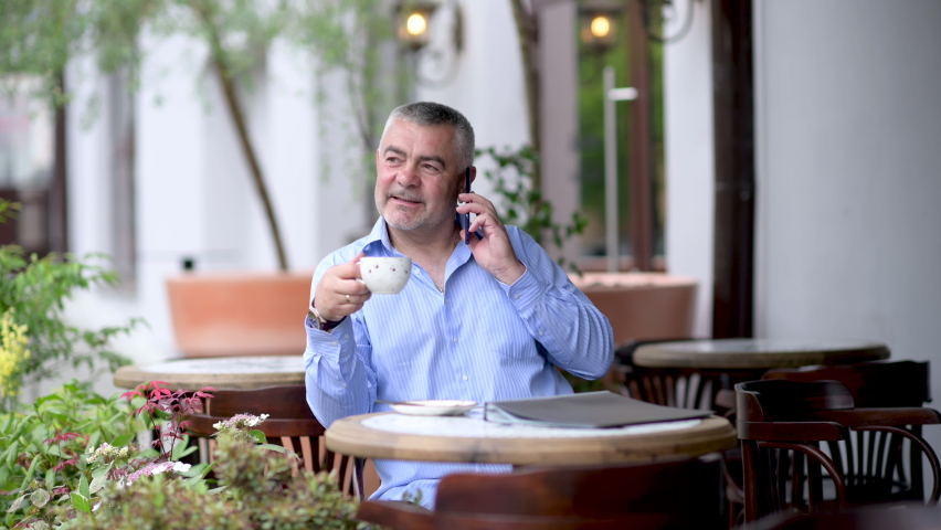Handsome caucasian middle aged businessman talking on the phone while sitting at the street cafe. | Shutterstock HD Video #1094345143