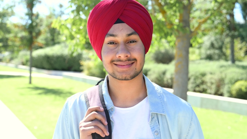 Young handsome Indian man in red traditional turban and casual shirt standing outdoors, smiling pleasant student with backpack looking at the camera, headshot