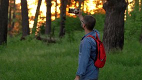 Male tourist with backpack make photos and shoot video on phone in summer forest at sunset. Travel blogger