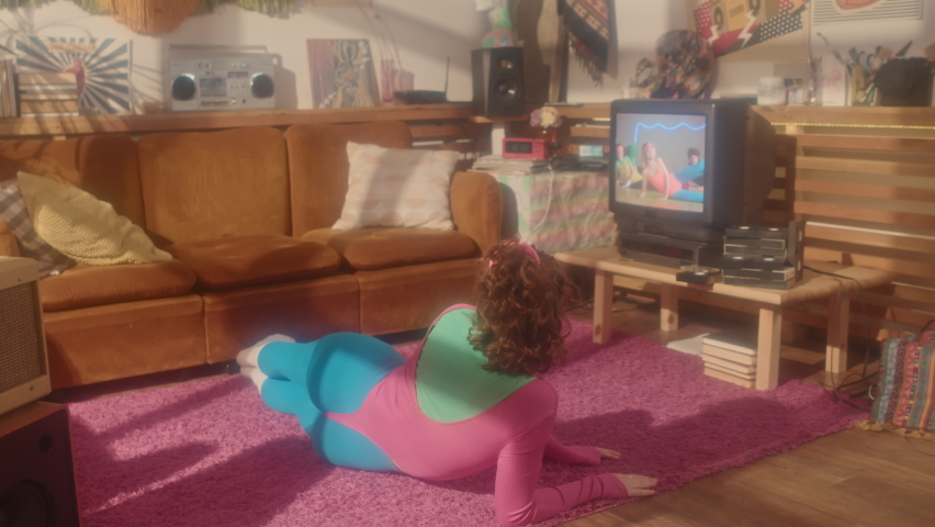 Wide rear of red-haired girl in eighties aerobic outfit exercising in living room in afternoon, watching tape on retro TV set Royalty-Free Stock Footage #1094354939