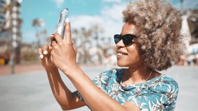 Cheerful African grey haired woman wearing sunglasses filming video of street by mobile outdoors