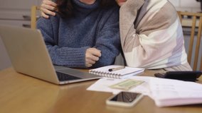 unrecognizable female couple sitting wrapped in blanket holding hands watching laptop screen. kitchen table smartphone papers work calculator, women cold hugging warming together, talking video call. 