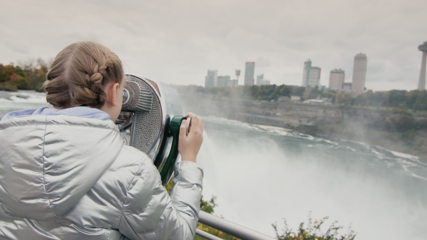 The girl looks through stationary binoculars at the beautiful landscape of Niagara Falls. Famous landmark on the border of the UNITED STATES and Canada