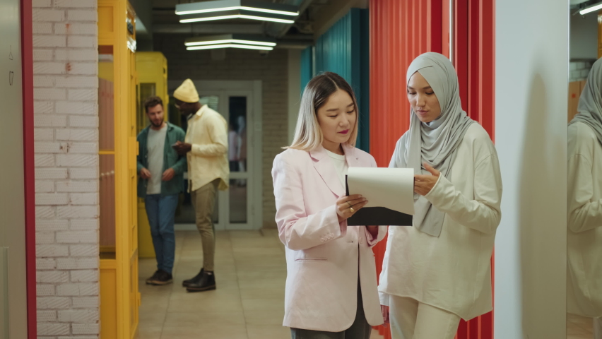 An Asian business woman and a Muslim business woman are discussing a project while standing in the corridor.Different Races,Diverse People,Creative Team,Business Partners