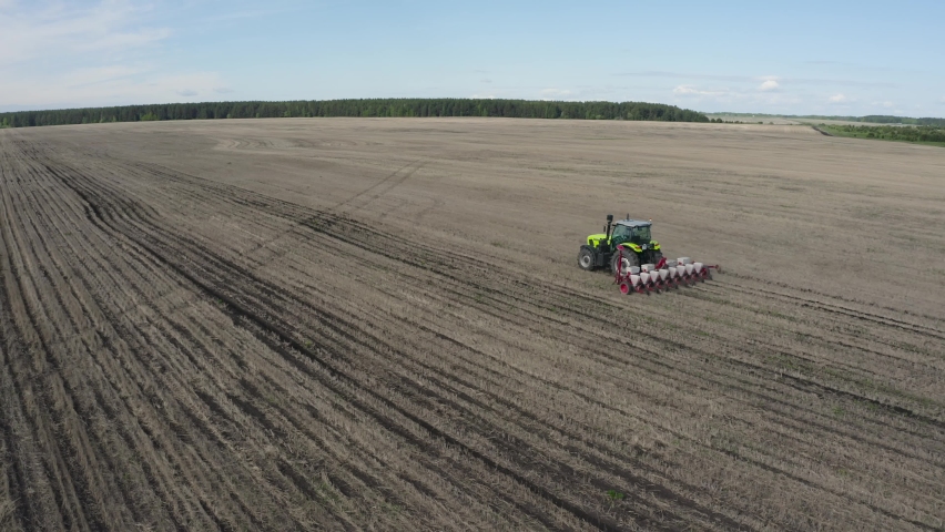 A green tractor plows an empty field. He rides, and furrows remain behind him. Forest is far away. Sky is blue. A bird flies over the equipment | Shutterstock HD Video #1094387915