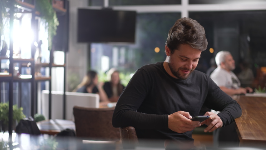Happy young male customer sitting at coffee shop holding phone. Portrait of a smiling man using technology looking at camera Royalty-Free Stock Footage #1094391417