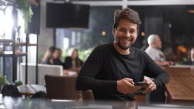 Happy young male customer sitting at coffee shop holding phone. Portrait of a smiling man using technology looking at camera - Powered by Shutterstock - Get 15% off with code: PIKWIZARD15