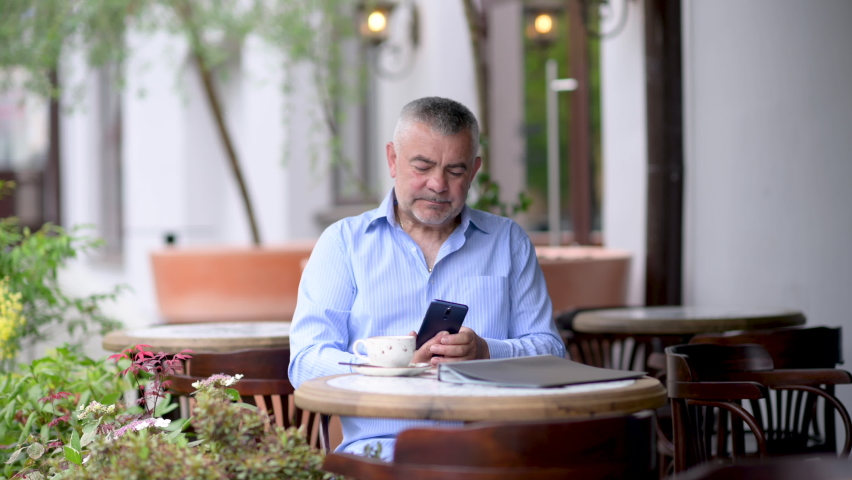 Handsome caucasian middle aged businessman sitting at the street cafe uses phone texting scrolling tapping. Man sitting at table in street cafe enjoying morning coffee. | Shutterstock HD Video #1094391809