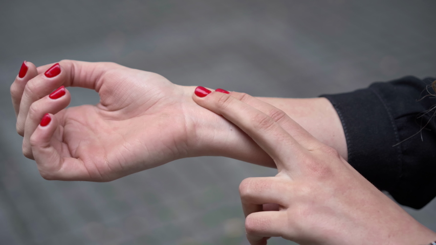 Close-up female hand taking pulse on wrist with fingers outdoors. Unrecognizable slim young Caucasian woman standing on park alley pavement. Anxiety and panic concept