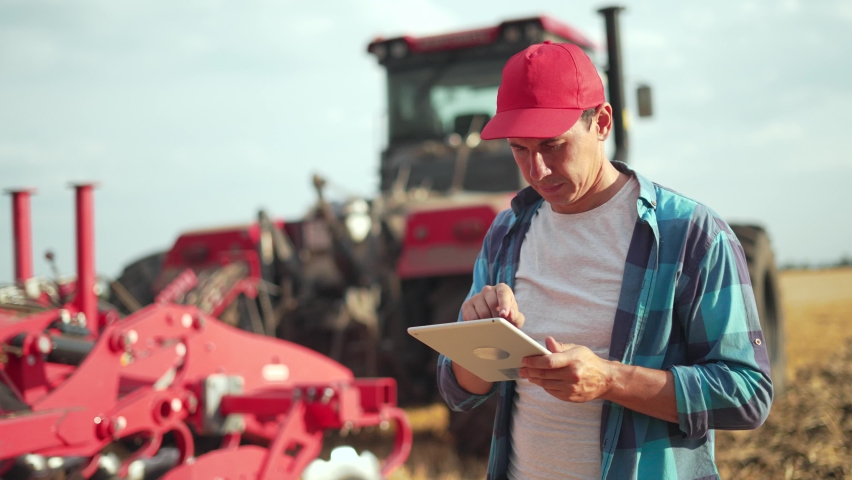 Farmer works with tablet in wheat field. work of tractor on plantation. Agriculture. Digital tablet in hands of agronomist farmer. Tractor cultivates wheat field. work of an agronomist in field. Royalty-Free Stock Footage #1094397311