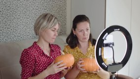 two female bloggers shoot video for blog hold pumpkins in hands talk about happy halloween or thanksgiving day. female couple streaming video webinar on smartphone camera in circular lamp tripod 