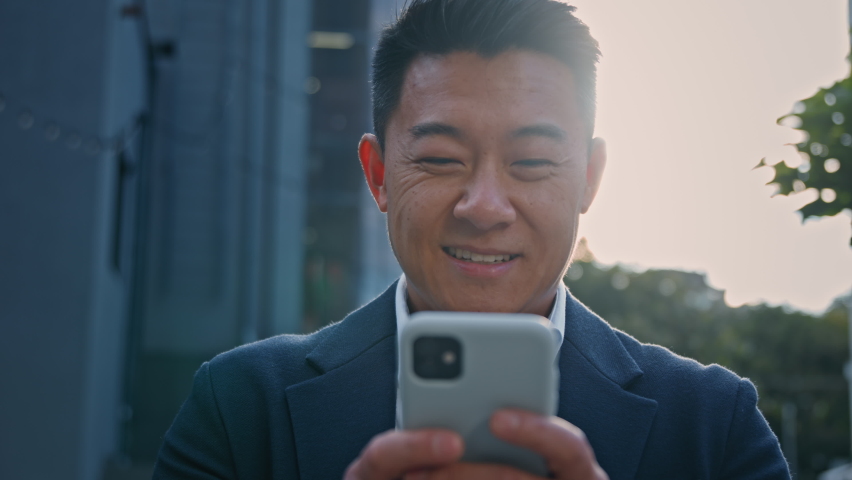 Close up smiling happy adult Asian Korean 40s businessman in city outdoors in sun light sunbeams background using smartphone with smile chatting in social network man texting mobile phone web online Royalty-Free Stock Footage #1094413985