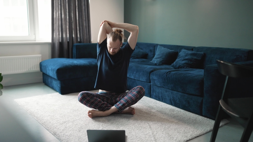 Positive blond man wearing blue t-shirt doing yoga stretching exercises on the floor at home