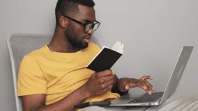 African american man in glasses looking in a notebook student and working in a laptop checking his knowledge at home online quarantine online training in a yellow t-shirt