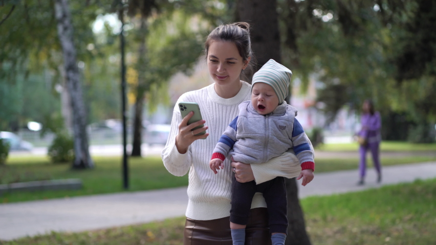 A mother with a child in her arms walking along the path is typing a message on the phone or browsing the news feed