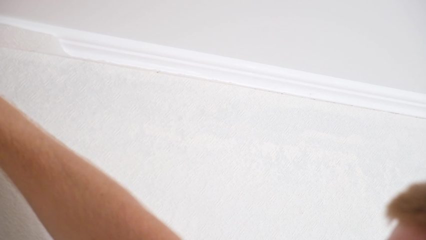 A man cuts wallpaper near ceiling moldings using box cutter and putty blade. Wallpapering. Repair.