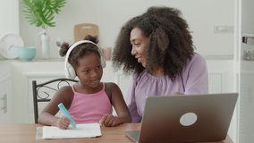 Pleasant African American young woman, carrying and loving mom helps her little daughter with study online. They are watching a video lesson together on laptop, sitting in a cozy light home interior