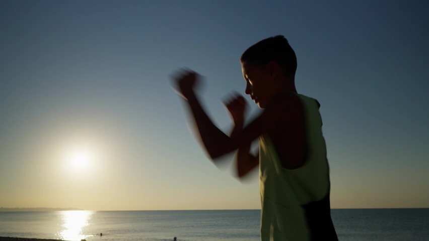 Young male boxer or karate fighter training on the beach during sunrise. Silhouette shot of boy doing shadow boxing in the morning. High quality 4k footage