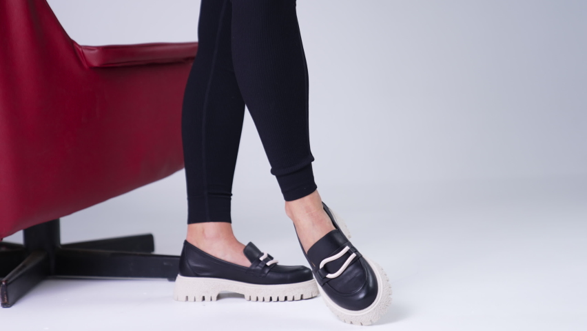 Unrecognized female in black pants wears modern shoes on white soles. Woman standing beside red chair demonstrating stylish footwear. White backdrop. | Shutterstock HD Video #1094449917
