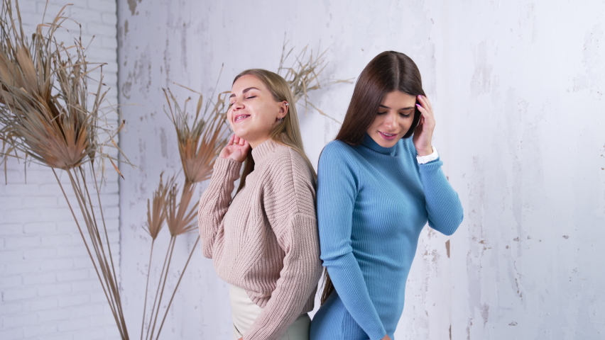 Two female models in warm fashionable clothes standing back to back. Ladies posing for camera in studio. White wall backdrop.